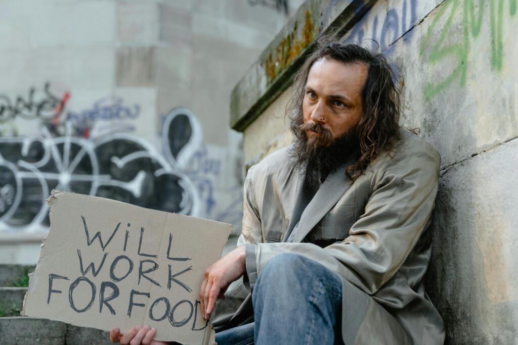 Man holding a “Will work for food” sign during an economic downturn, illustrating why learning how to survive an economic crash matters.